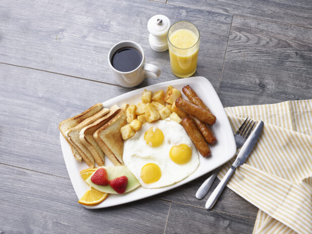 3 eggs sunny side up, breakfast sausages, breakfast potatoes, white toast, and fruit on a white platter alongside coffee and juice on a gray wooden background
