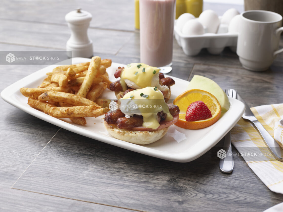 A breakfast plate of eggs sausage benedict and seasoned fries with fruit garnish and a smoothie in a glass