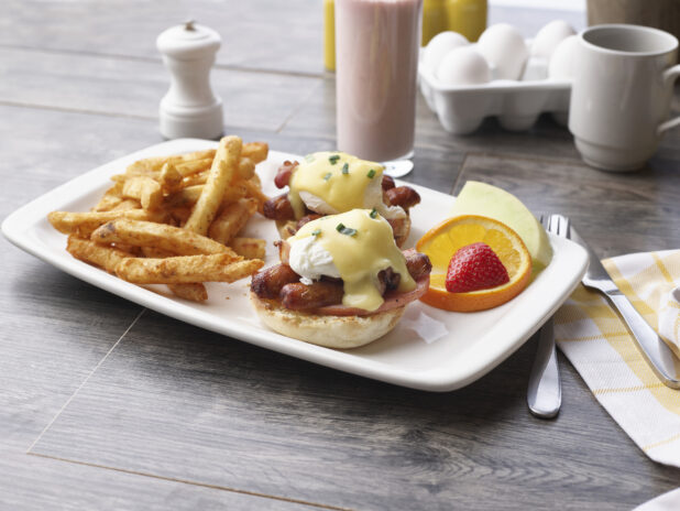 A breakfast plate of eggs sausage benedict and seasoned fries with fruit garnish and a smoothie in a glass