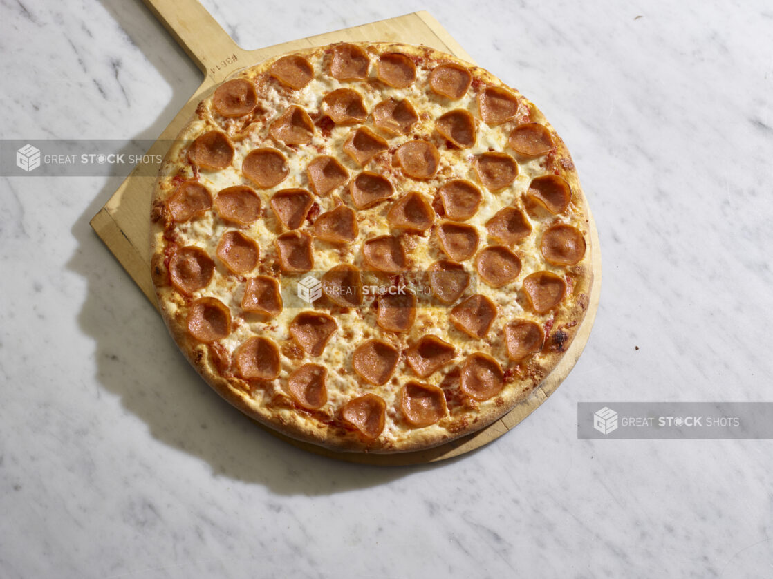 A traditional style pepperoni pizza on a wooden pizza peel on a marble background