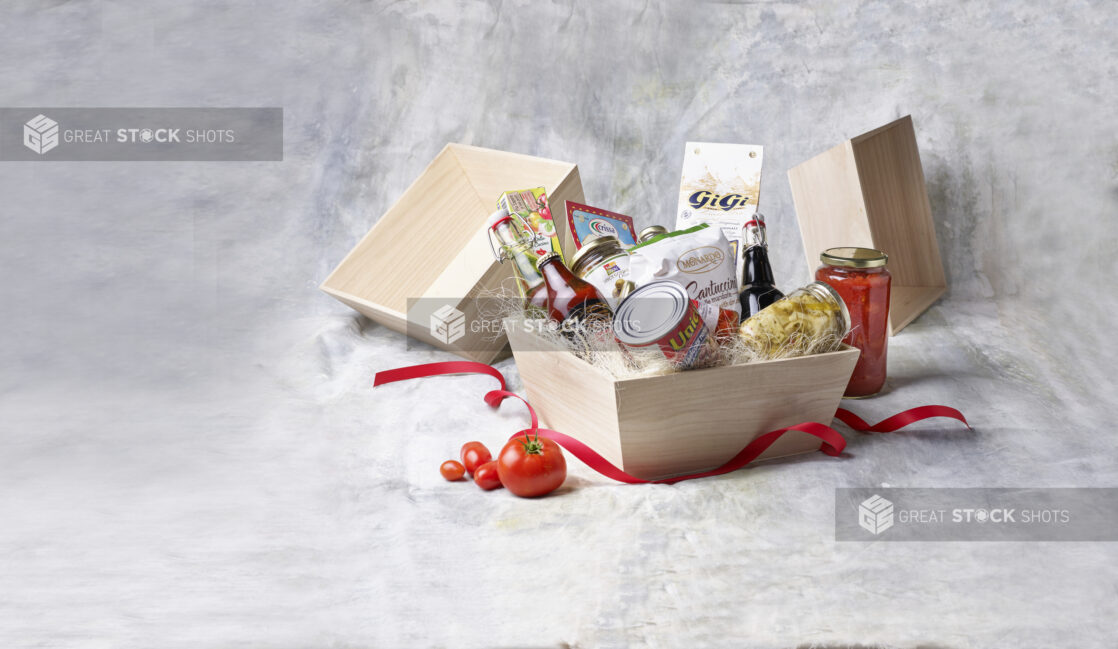 Deep Square Wood Tray Gift Basket Filled with Italian Pantry Products, Jars and Canned Goods with a Red Ribbon Against a Canvas Background