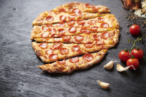 Close Up of a Sliced Whole Pepperoni Pizza Surrounded by Fresh Ingredients on a Black Painted Wood Surface