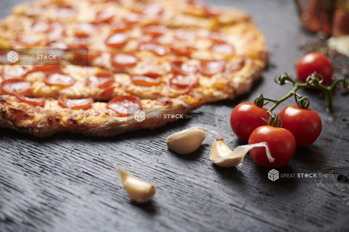 Close Up of Raw Garlic Cloves and Cherry Tomatoes with Sliced Whole Pepperoni Pizza in the Background on a Black Painted Wood Surface