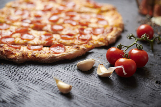 Close Up of Raw Garlic Cloves and Cherry Tomatoes with Sliced Whole Pepperoni Pizza in the Background on a Black Painted Wood Surface