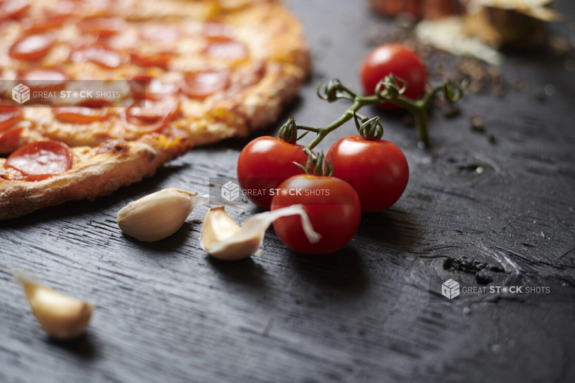 Close up of Cherry Tomatoes on the Vine with Raw Garlic Cloves and a Sliced Whole Pepperoni Pizza on a Black Painted Wood Surface