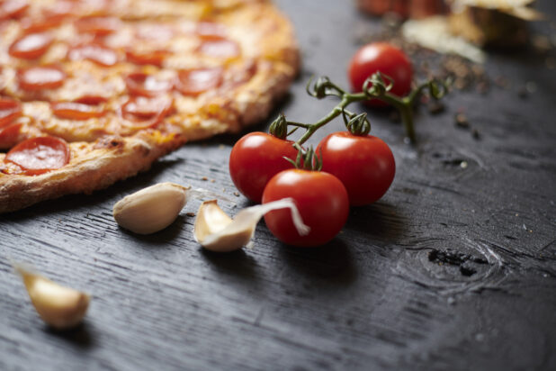 Close up of Cherry Tomatoes on the Vine with Raw Garlic Cloves and a Sliced Whole Pepperoni Pizza on a Black Painted Wood Surface