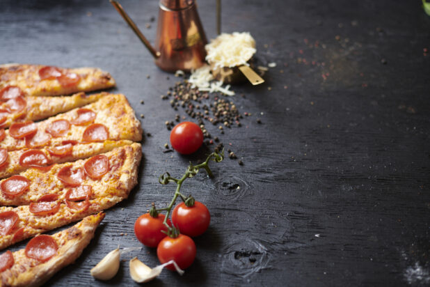 Thin strips of pepperoni pizza surrounded by aromatic ingredients on a black wooden background