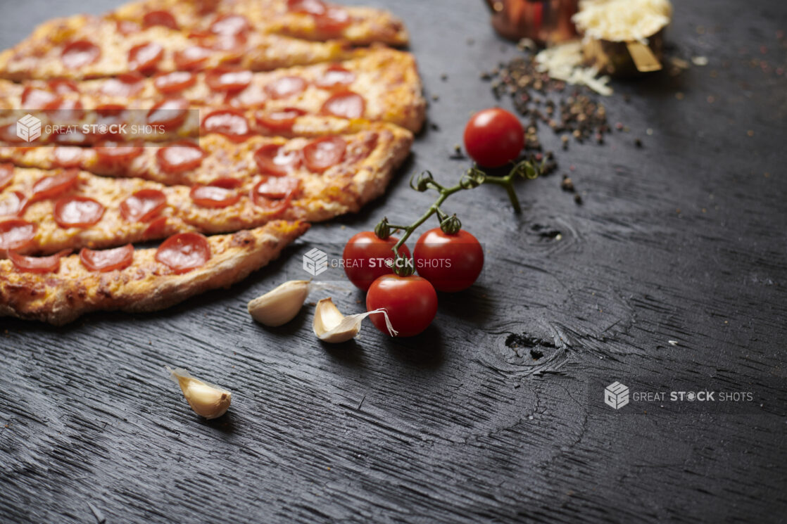 Fresh Cherry Tomatoes on the Vine with Raw Garlic Cloves and a Sliced Whole Pepperoni Pizza on a Black Painted Wood Surface