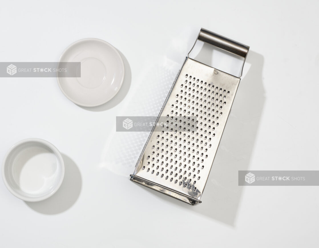 Overhead View of Stainless Steel Cheese Grater and Round White Ceramic Dishes, on a White Background for Isolation