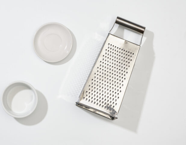 Overhead View of Stainless Steel Cheese Grater and Round White Ceramic Dishes, on a White Background for Isolation