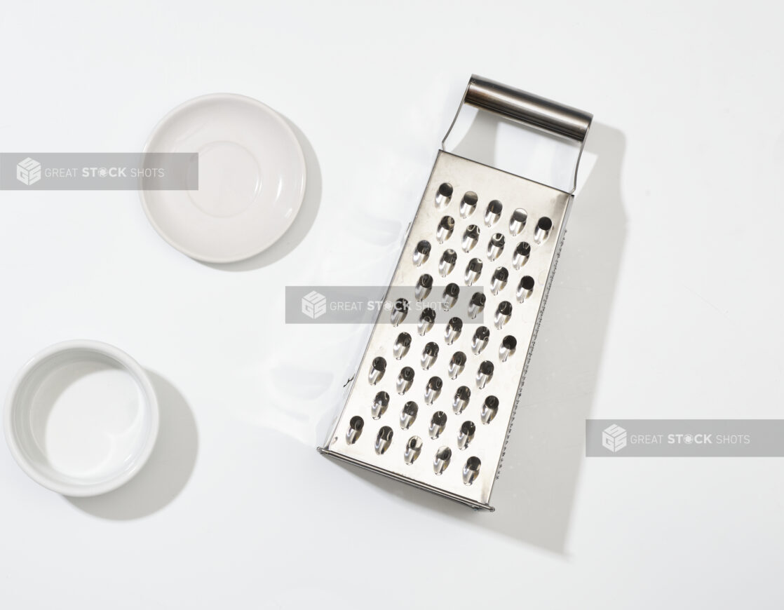 Overhead View of Stainless Steel Cheese Grater and Round White Ceramic Dishes, on a White Background for Isolation - Variation