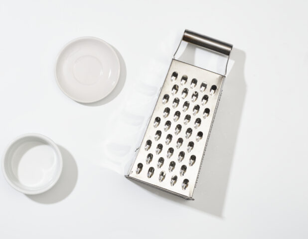 Overhead View of Stainless Steel Cheese Grater and Round White Ceramic Dishes, on a White Background for Isolation - Variation