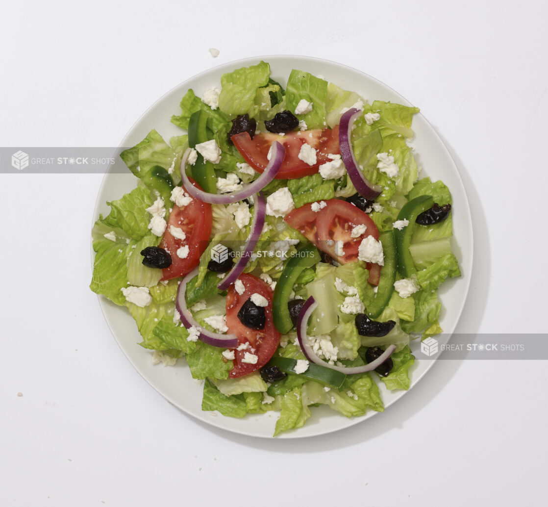 Overhead View of a Greek Salad with Sliced Tomatoes, Green Peppers, Red Onions, Black Olives and Feta Cheese on a Round White Ceramic Dish, on a White Background for Isolation