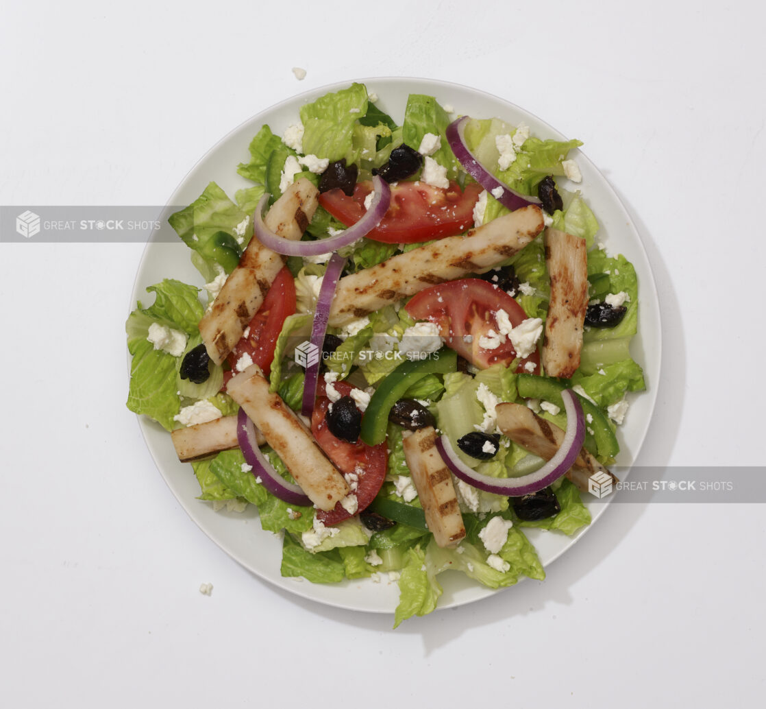 Overhead View of a Greek Salad with Sliced Tomatoes, Green Peppers, Red Onions, Black Olives, Feta Cheese and Grilled Chicken on a Round White Ceramic Dish, on a White Background for Isolation