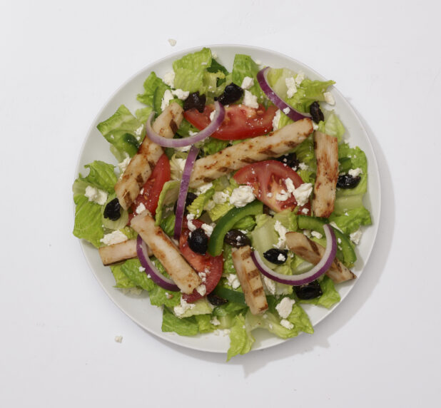Overhead View of a Greek Salad with Sliced Tomatoes, Green Peppers, Red Onions, Black Olives, Feta Cheese and Grilled Chicken on a Round White Ceramic Dish, on a White Background for Isolation