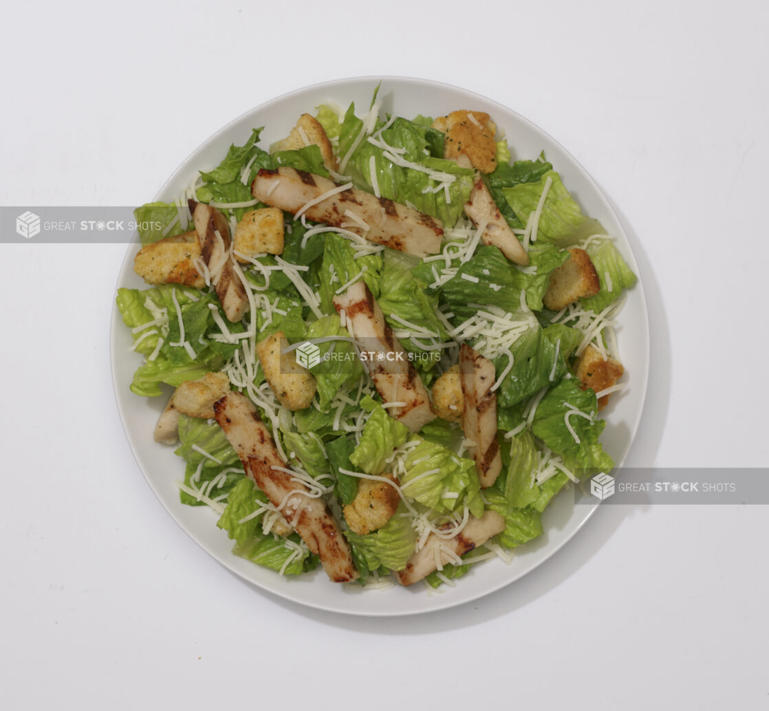 Overhead View of a Caesar Salad with Shredded Parmesan, Croutons and Grilled Chicken on a Round White Ceramic Dish, on a White Background for Isolation