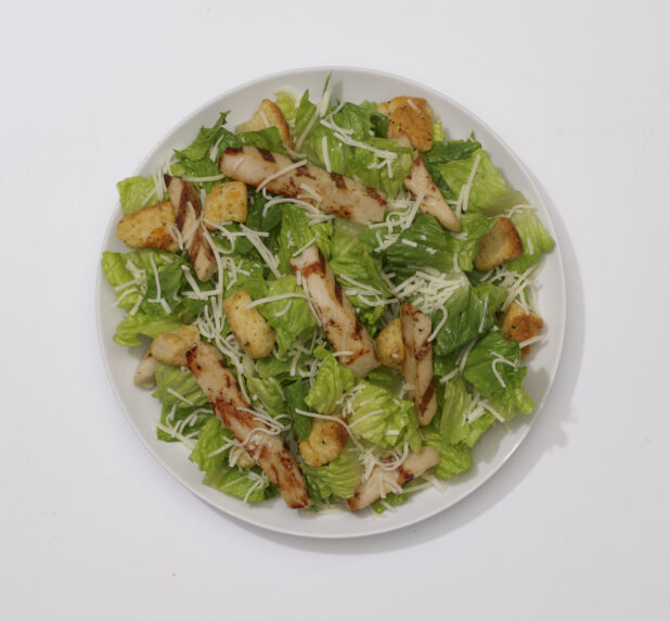 Overhead View of a Caesar Salad with Shredded Parmesan, Croutons and Grilled Chicken on a Round White Ceramic Dish, on a White Background for Isolation