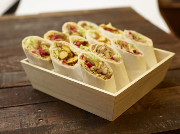 Deep Square Wood Serving Tray Filled with Chicken Shawarma, BBQ Chicken and Falafel Pita Wraps with Fresh Vegetable Toppings, Rolled in Parchment Paper on a Dark Wooden Table Surface