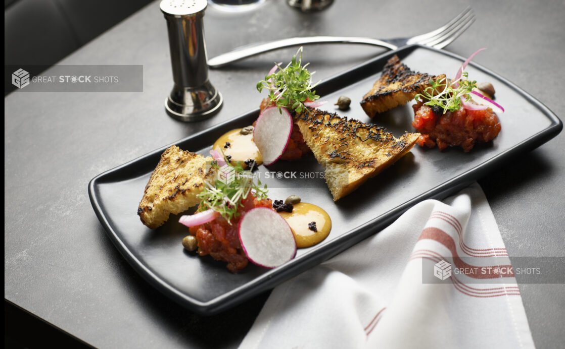 An elegant Salmon Crudo appetizer on a dark plate with toast and microgreens