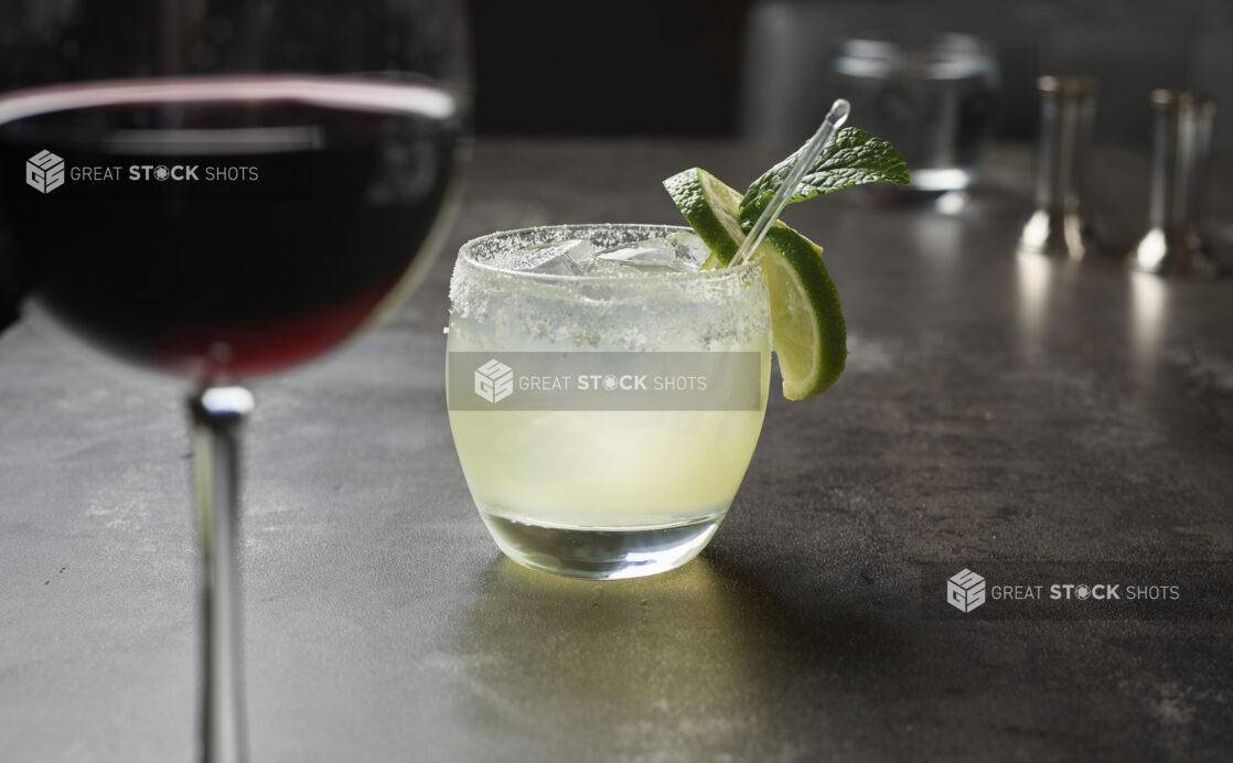 A margarita cocktail with lime garnish and fresh mint, glass of red wine in foreground on grey tabletop, close up, low angle