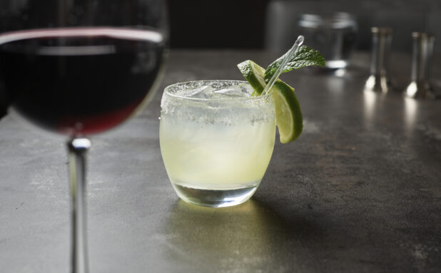 A margarita cocktail with lime garnish and fresh mint, glass of red wine in foreground on grey tabletop, close up, low angle