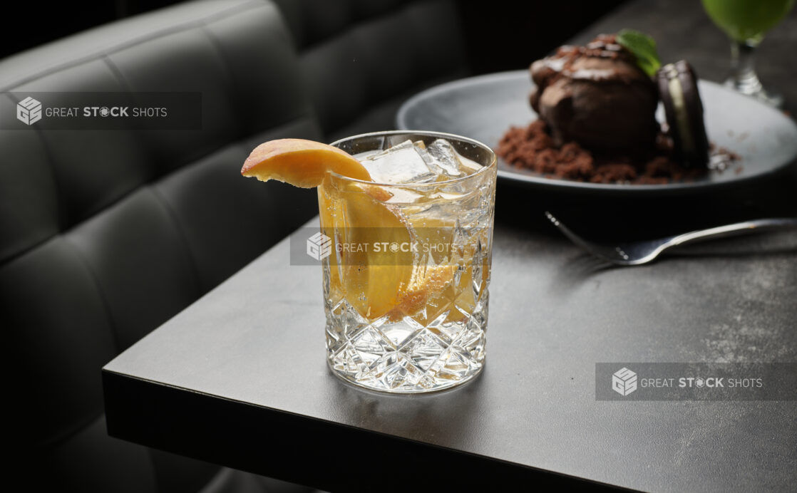 A cocktail in a crystal rocks glass with a peach slice garnish and ice cubes on a dark table, chocolate dessert in background