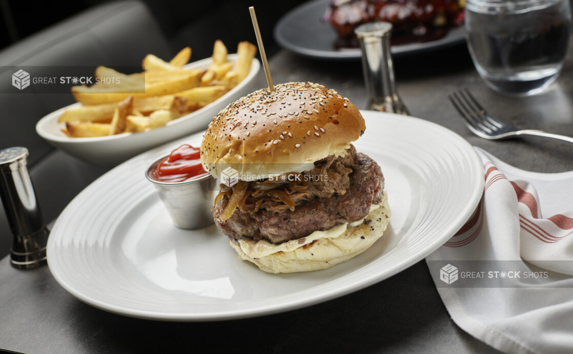 A beef burger on a brioche bun with caramelized onions and Dijon aioli on a white plate ketchup and a side of fries