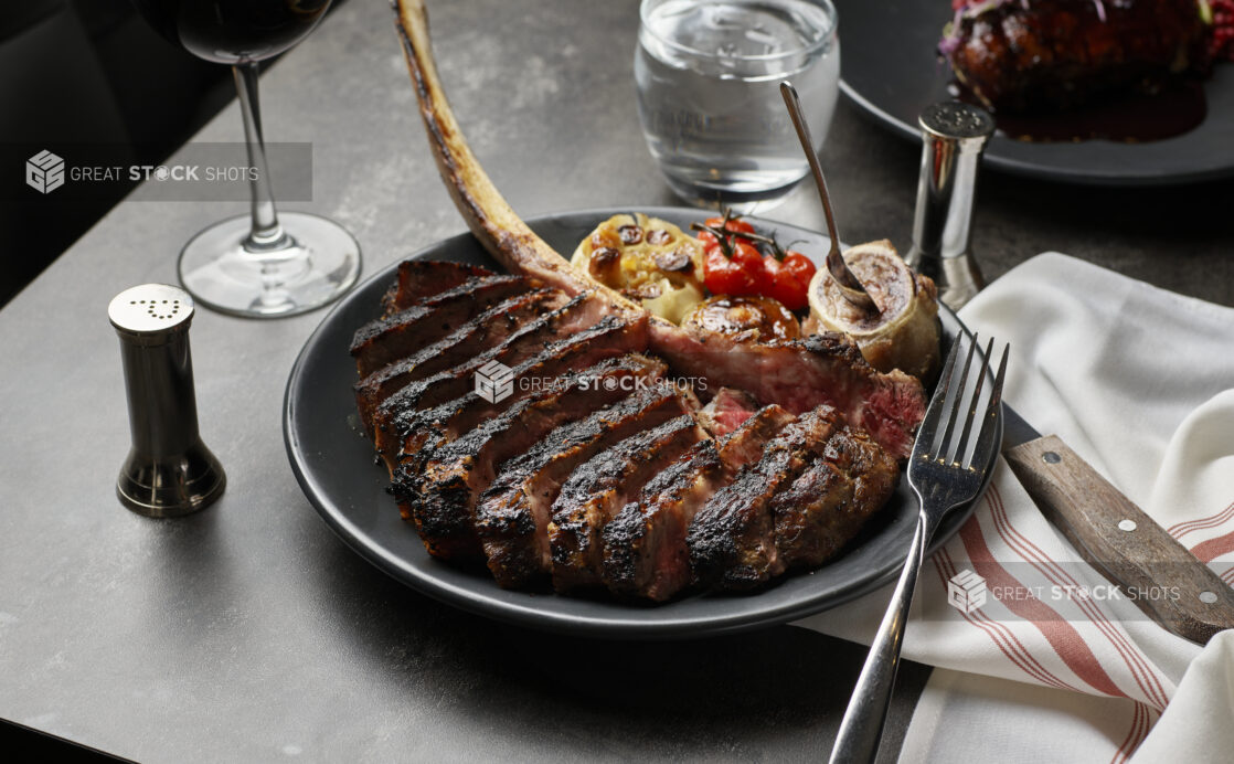 Sliced Tomahawk steak with roasted garlic, roasted mushroom, blistered cherry tomatoes, and marrow on a black plate