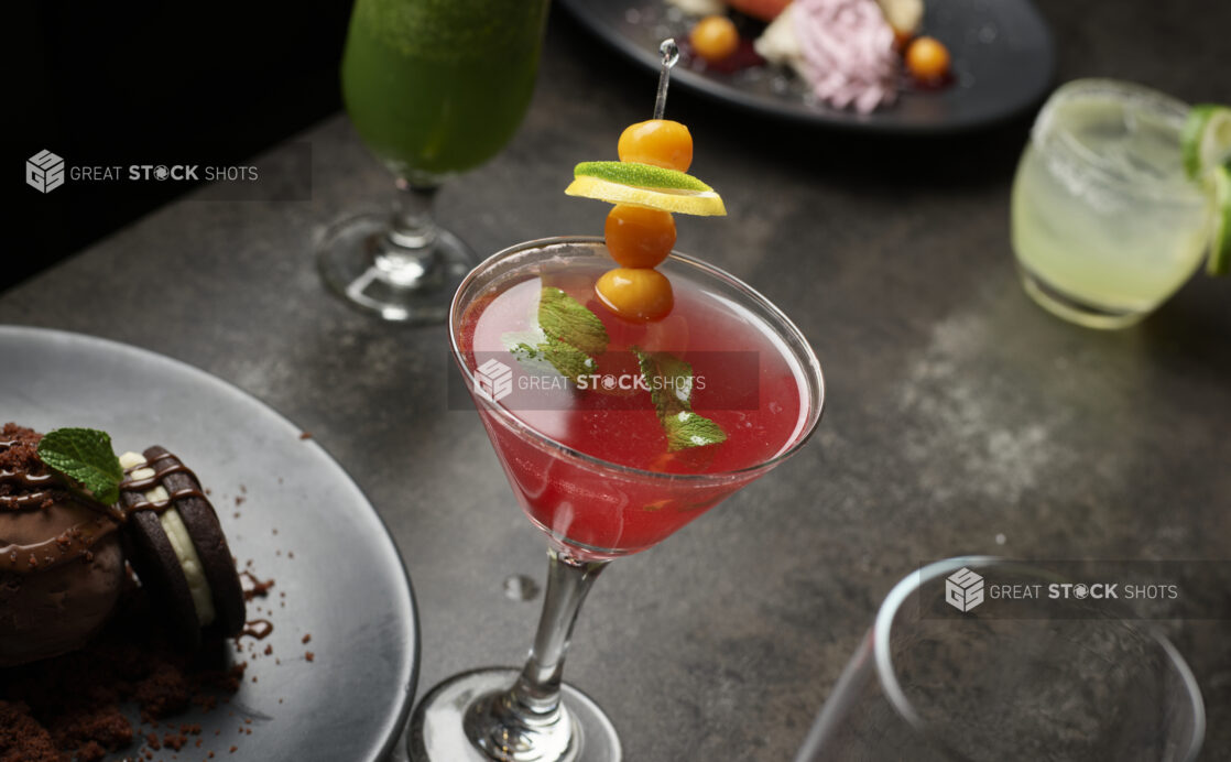 A red cocktail in a martini glass garnished with fresh mint, sea buckthorn and lime