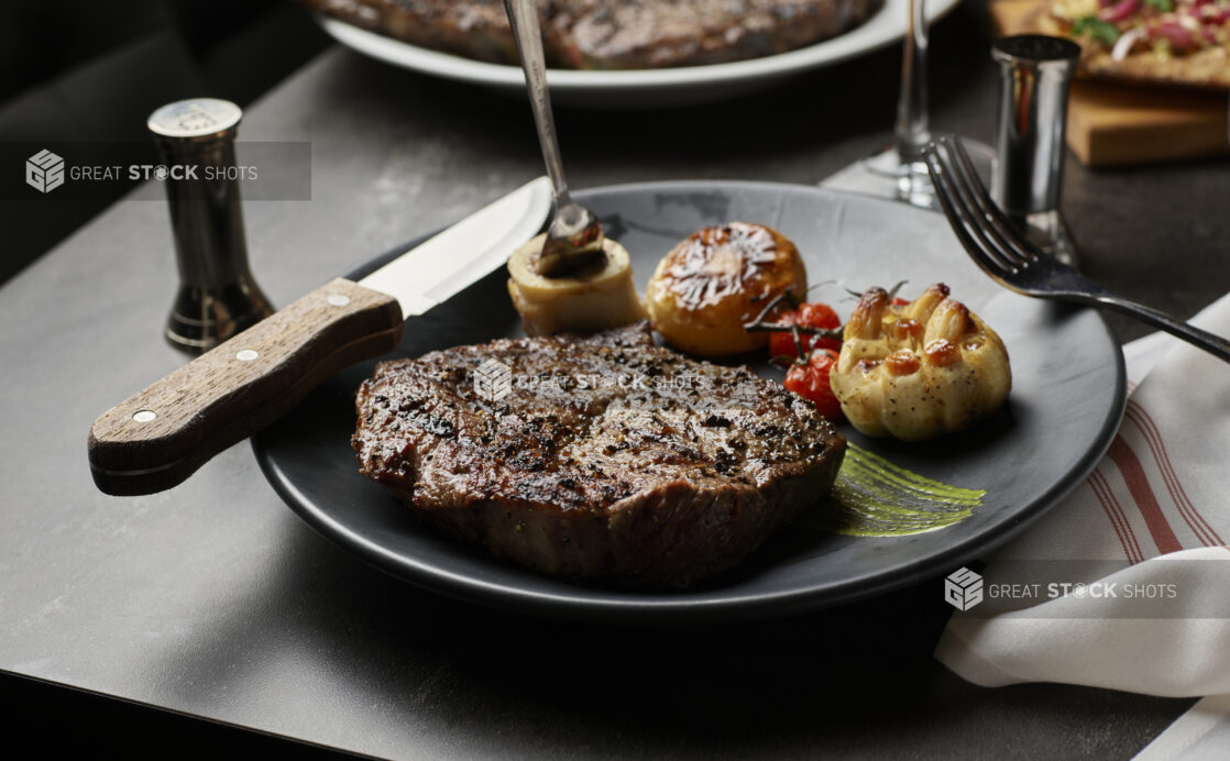 A boneless ribeye with roasted garlic blistered cherry tomatoes roasted mushroom and bone marrow on a black plate