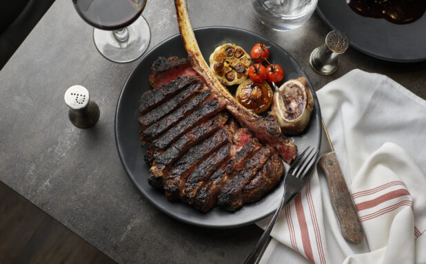 Overhead view of a sliced Tomahawk steak with roasted garlic, roasted mushroom, blistered cherry tomatoes, and marrow on a black plate