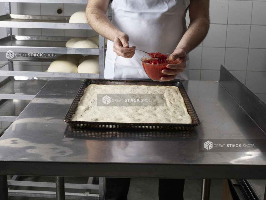 Pizza chef beginning to spread pizza sauce on a large uncooked square pizza