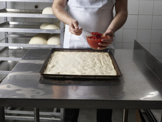 Pizza chef beginning to spread pizza sauce on a large uncooked square pizza