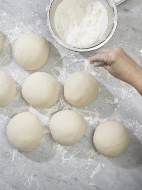 A baker sprinkling flour onto balls of pizza dough on a marble prep surface