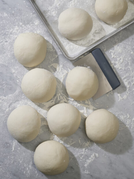 An overhead view of balls of raw pizza dough on a marble prep surface dusted with flour
