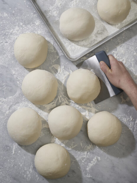 A baker lifting a ball of dough using a bench scraper with balls of pizza dough on a marble background
