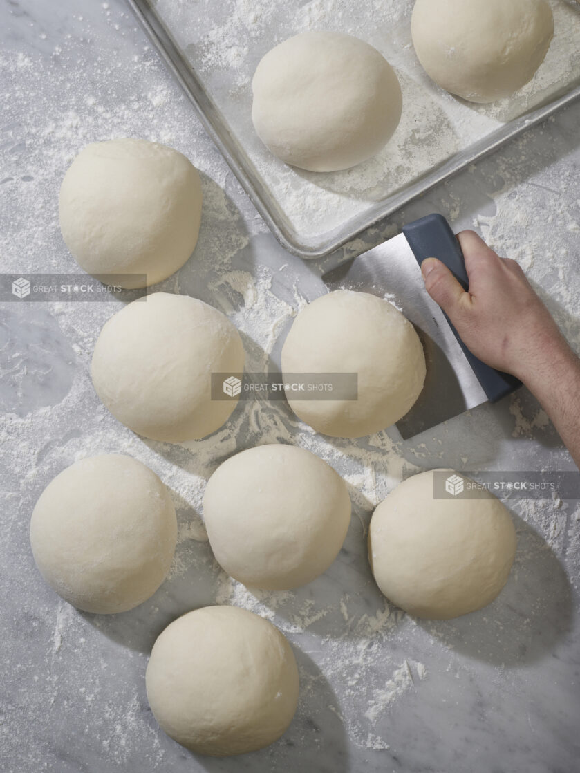 A baker lifting a ball of dough using a bench scraper with balls of pizza dough on a marble background