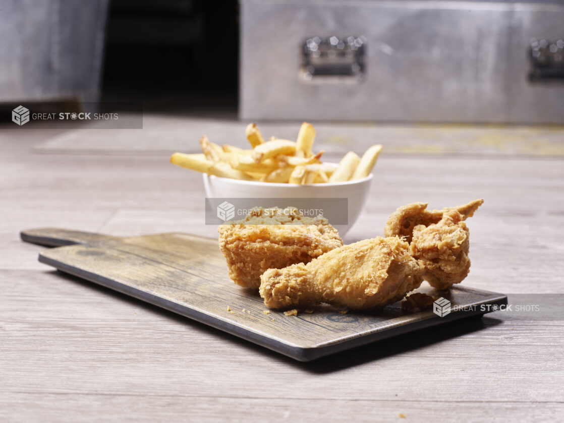 Fried chicken on a wooden board with a side of french fries in a small white ceramic bowl on a wooden background