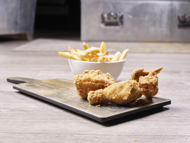 Fried chicken on a wooden board with a side of french fries in a small white ceramic bowl on a wooden background