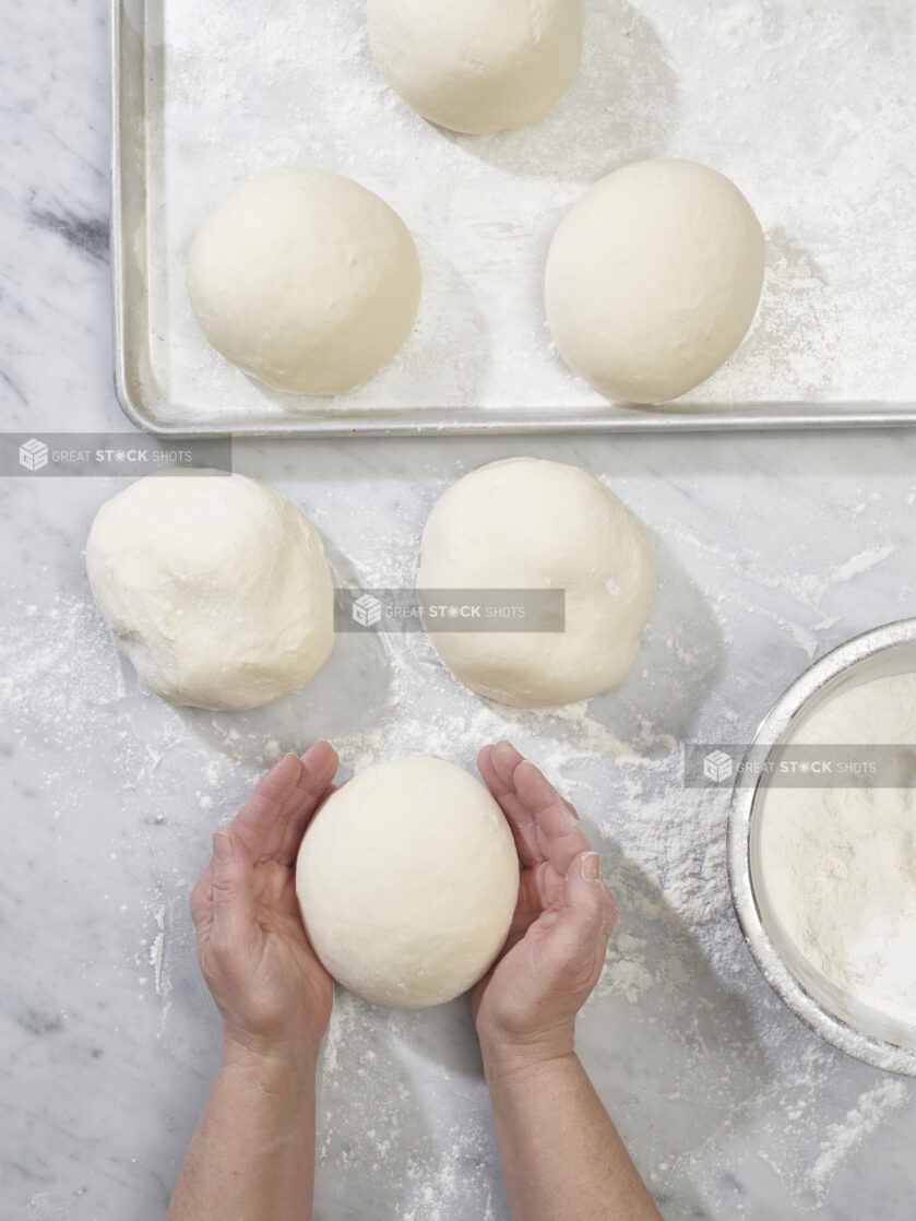 An overhead view of a baker's hands shaping raw pizza dough on a marble prep surface