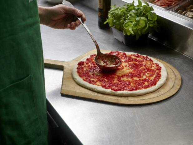 A Pizzaiolo spreading marinara sauce on stretched pizza dough in a commercial kitchen