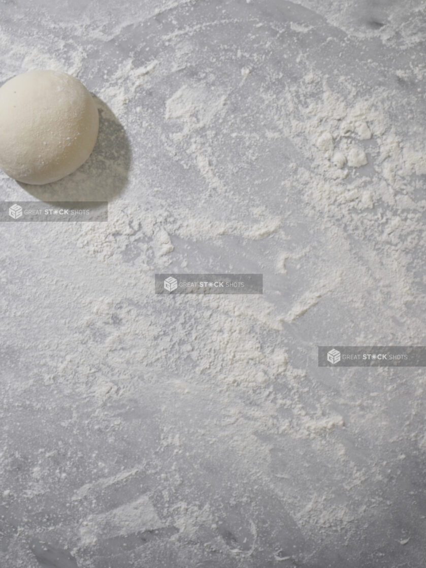 An overhead view of a ball of pizza dough on a marble background dusted with flour