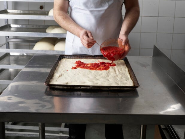 Pizza chef spreading pizza sauce on a square uncooked pizza in a commercial kitchen