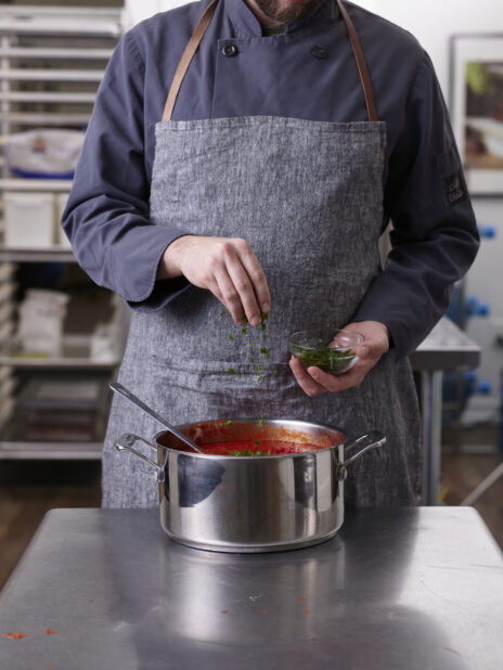 A chef with a gray apron standing over a pot of tomato sauce