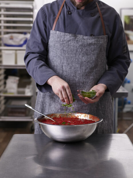 A chef in a gray apron seasoning tomato sauce in a steel bowl