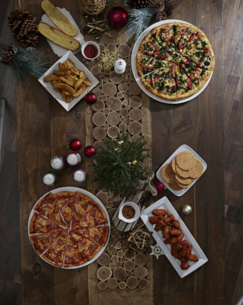 A dinner table decorated for Christmas with two large pizzas, a platter of wings, garlic bread, potato wedges and cookies, overhead view