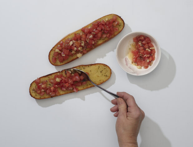 Chef's hand with a spoon adding bruschetta topping to a sliced garlic baguette