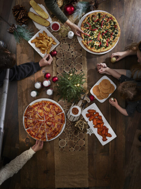 A family at a dinner table decorated for Christmas with two large pizzas, a platter of wings, garlic bread, potato wedges and cookies, overhead view