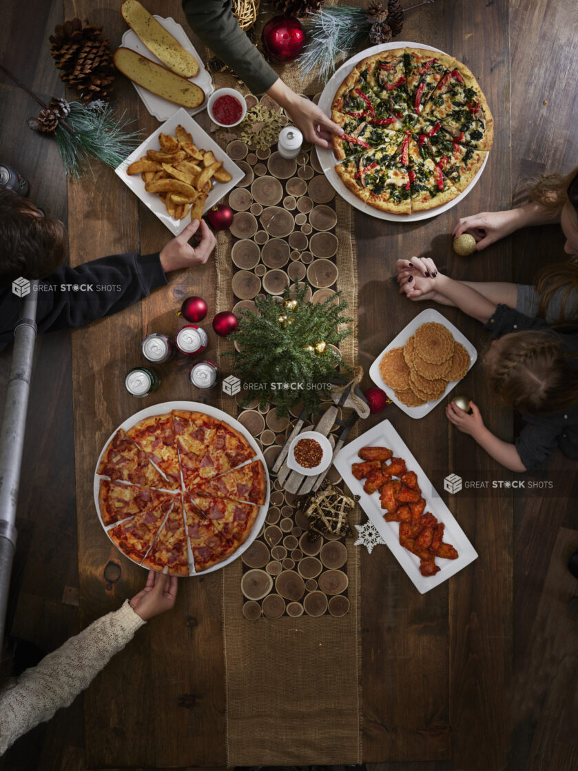 A family at a dinner table decorated for Christmas with two large pizzas, a platter of wings, garlic bread, potato wedges and cookies, overhead view