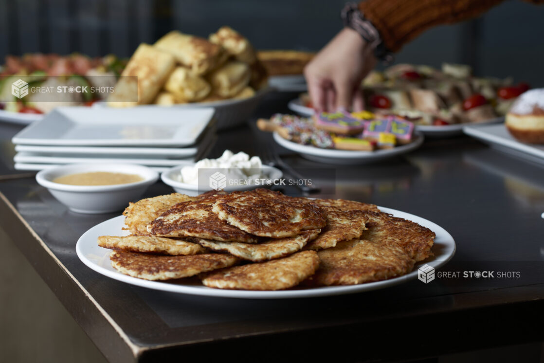 A plate of latkes on a buffet table of Hanukkah food with a hand reaching for cookies in the background